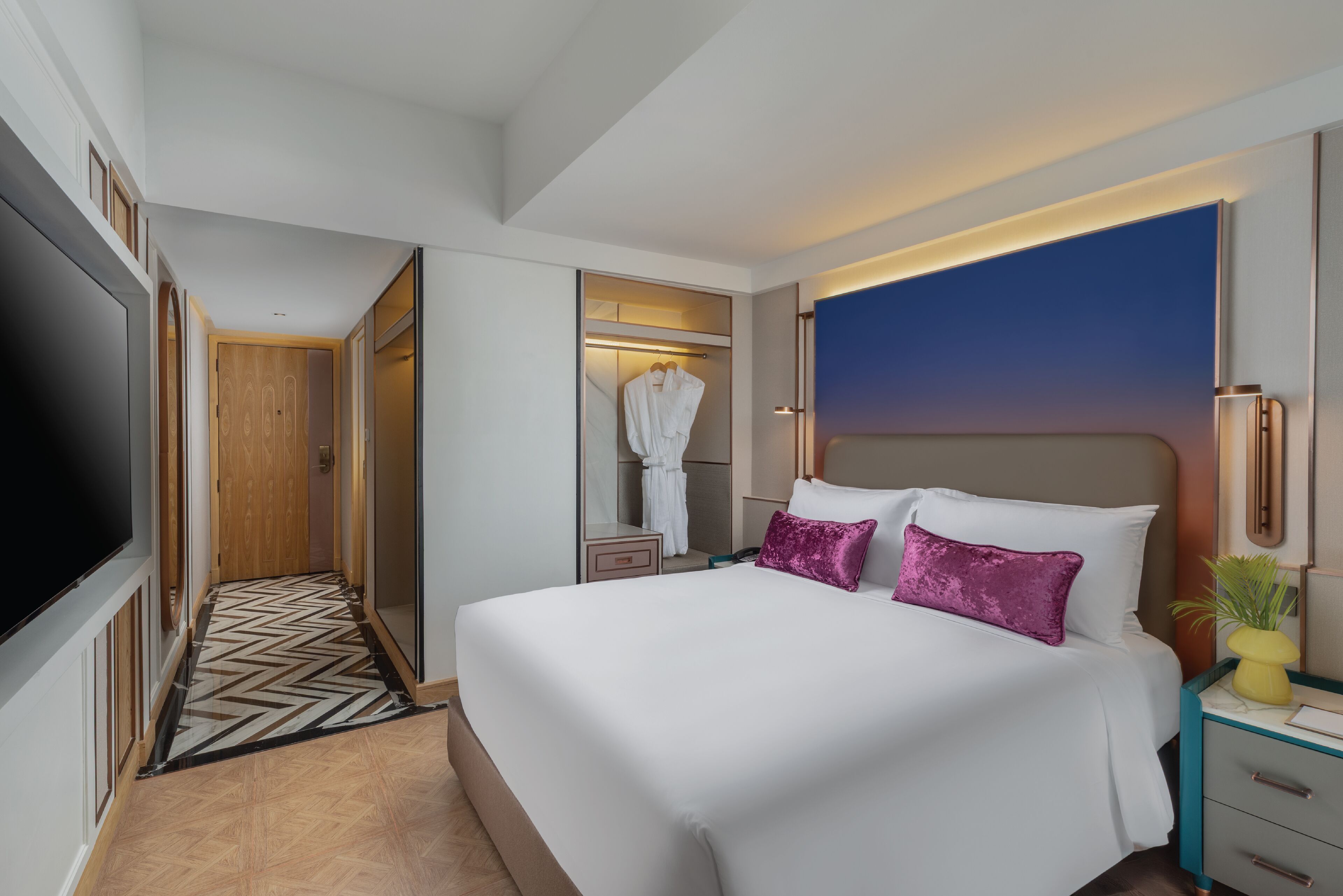 A hotel room with a large bed, a walk-in closet, a TV, and a bedside table with a lamp.