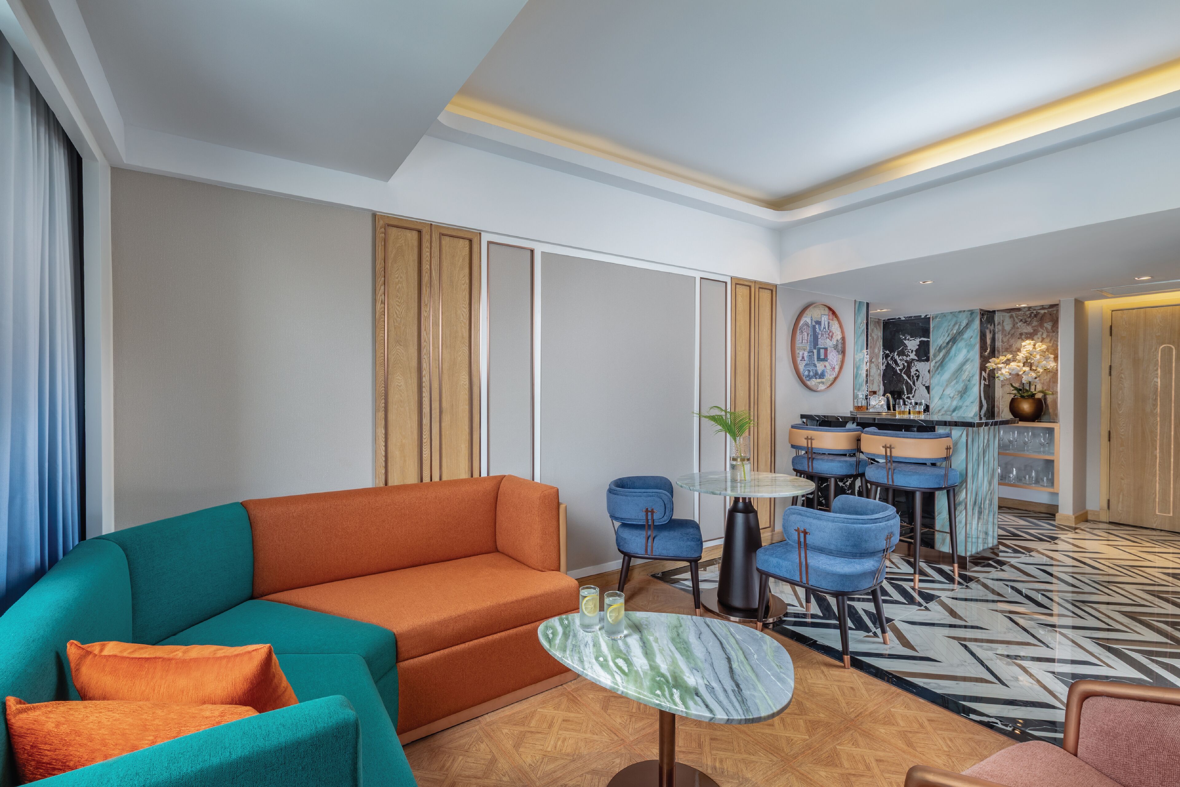 Una sala de estar moderna con un sofá verde azulado y naranja, una mesita redonda y una zona de bar con sillas azules.