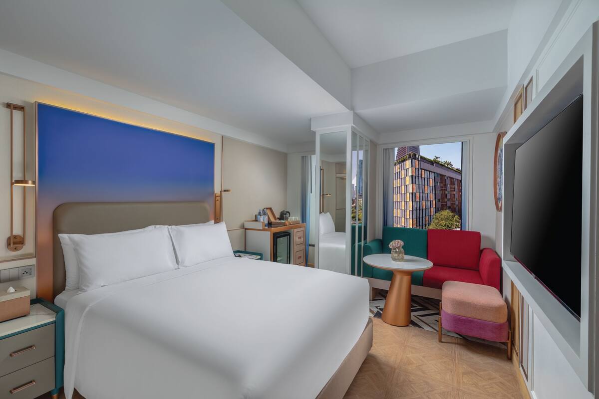 A hotel room with a large bed, a desk, a chair, a TV, and a balcony with a city view.