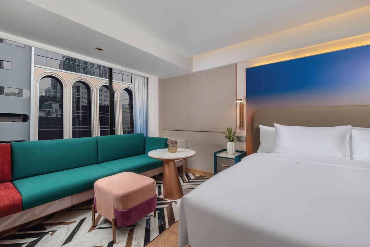 A modern hotel room with a teal sofa, a small round table, a bed with white linens, and a view of the city through large windows.