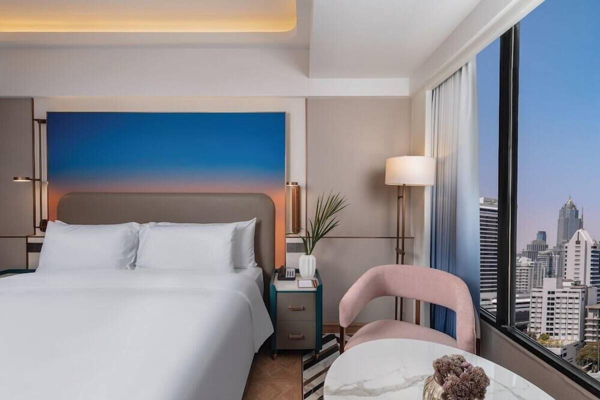 A hotel room with a large bed, a nightstand with a lamp, a chair, and a view of the cityscape.