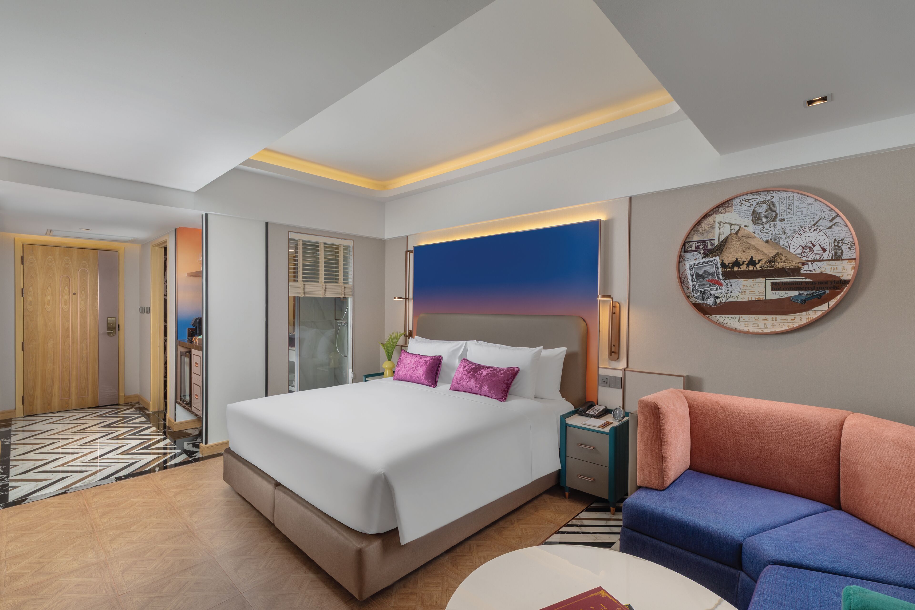 A modern hotel room with a large bed, a sofa, a round wall decoration, and a bathroom visible through an open door.