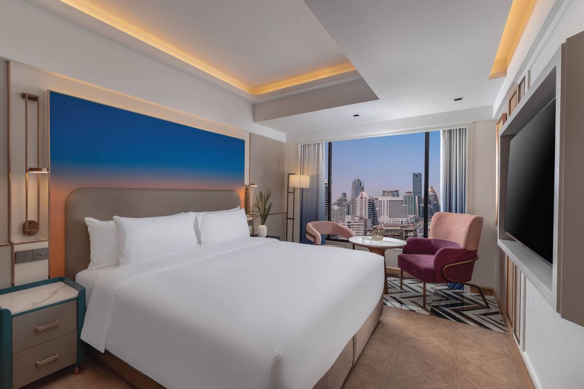 A hotel room with a large bed, a desk, a chair, a TV, and a city view.