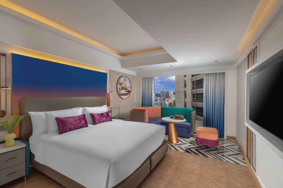A modern hotel room with a large bed, a TV, a city view, and a cozy seating area.