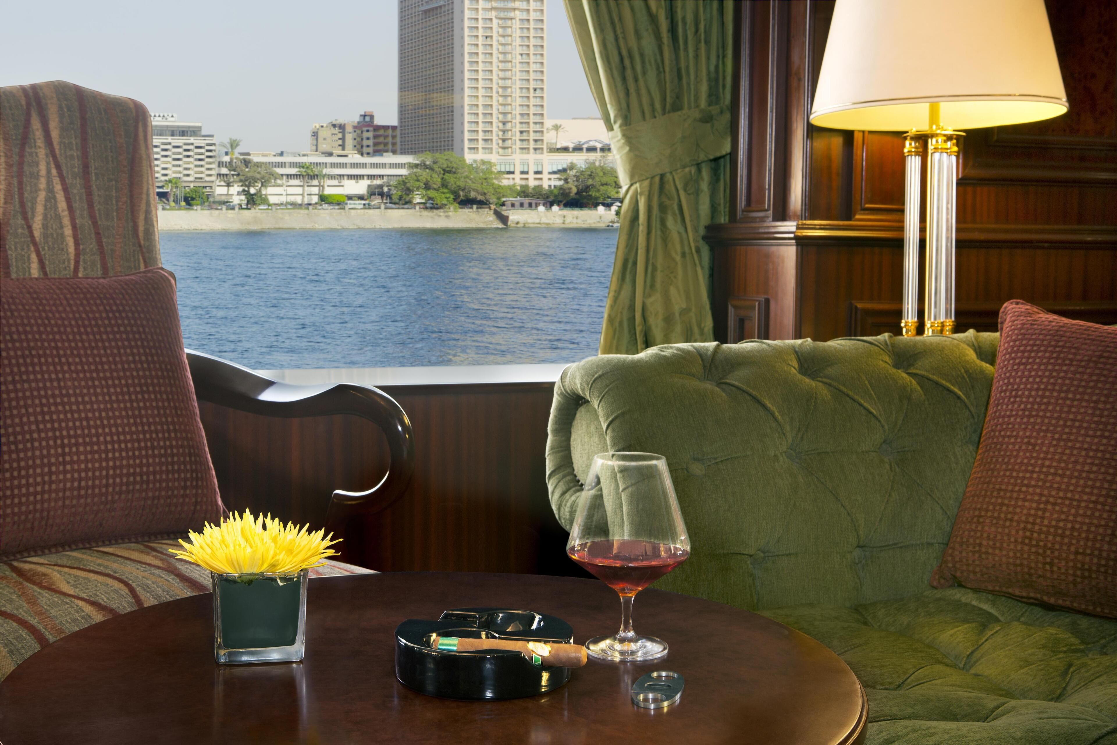 A room with a view of a body of water, a green upholstered sofa, a table with a glass of red wine, and a lamp.