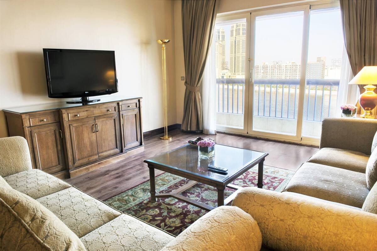 A living room with a flat-screen TV, wooden cabinet, sofa, coffee table, and a view of a cityscape through a balcony.