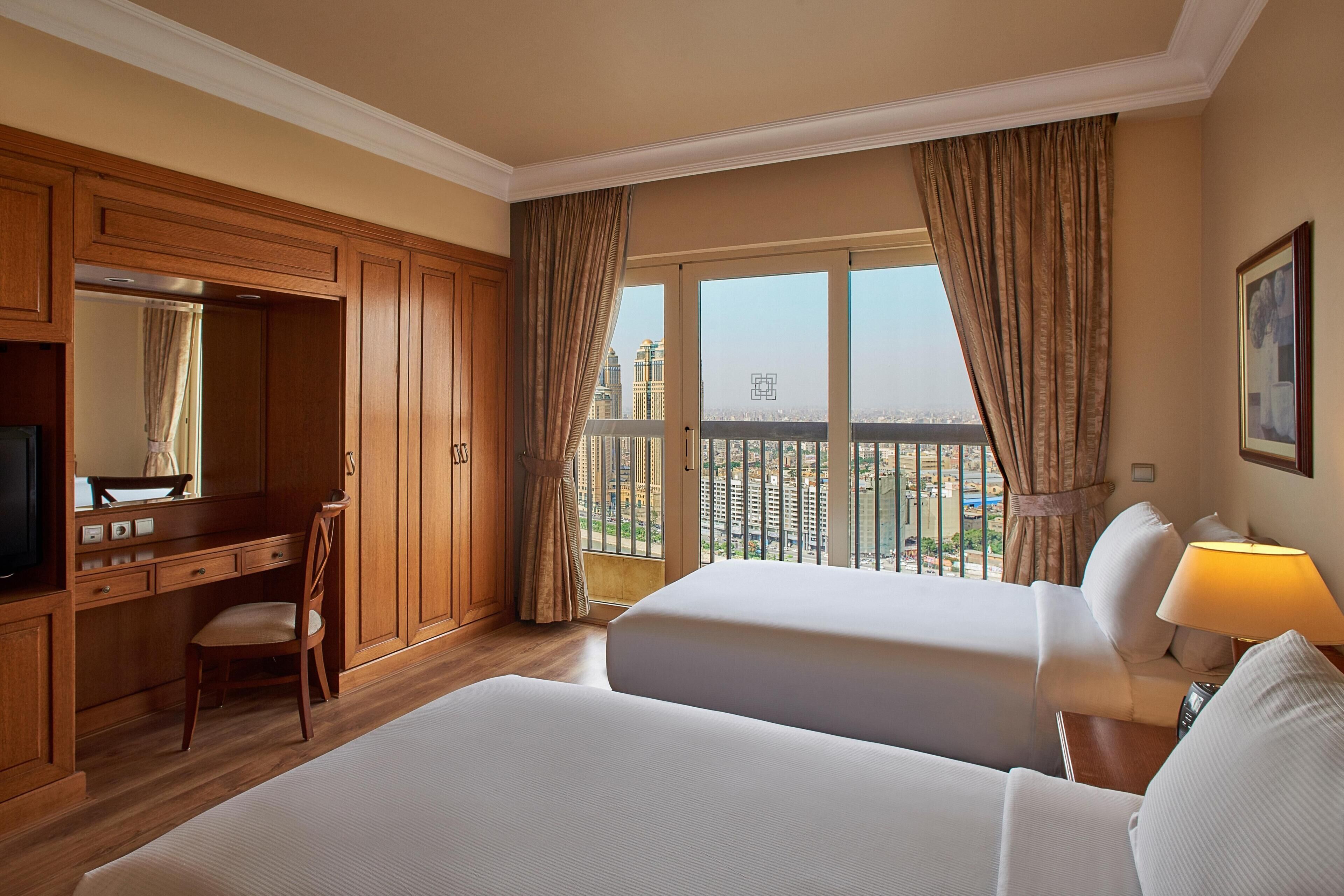 A hotel room with two beds, a desk, a chair, a TV, and a large window with curtains.