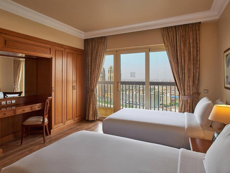 A hotel room with two beds, a desk, a chair, a TV, and a large window with curtains.