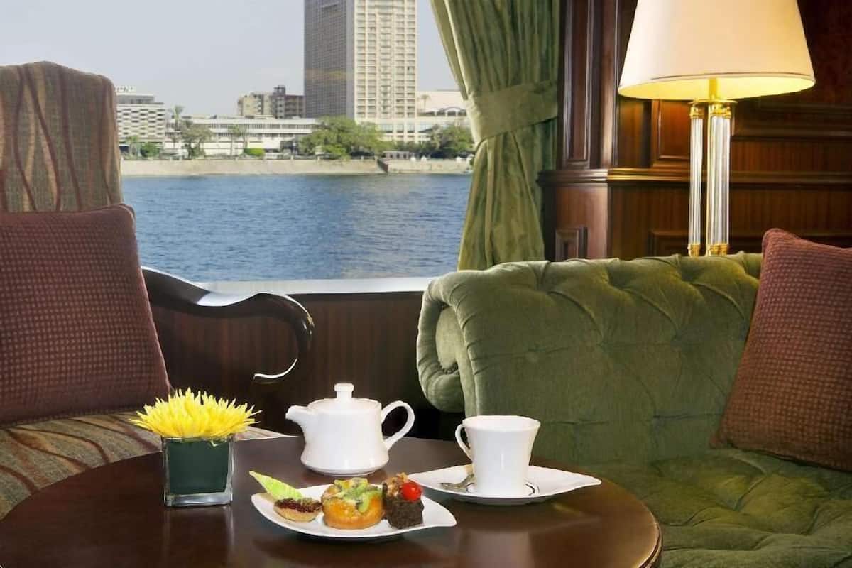 A hotel room with a view of a body of water and buildings, a green upholstered sofa, a small table with a teapot, a cup, and a plate of pastries.