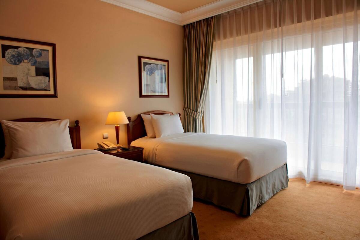 A hotel room with two beds, a nightstand with a lamp, a telephone, and two framed pictures on the wall.