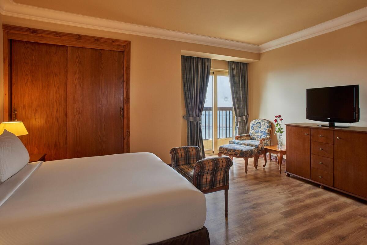 A hotel room with a large bed, a television, a wooden wardrobe, and a seating area with a chair and a small table.