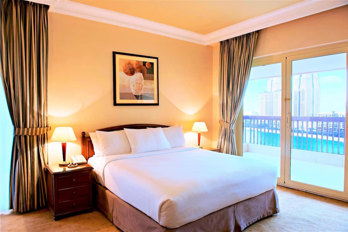 A hotel room with a bed, a nightstand with a lamp, a framed picture on the wall, and a view of a cityscape through a balcony.