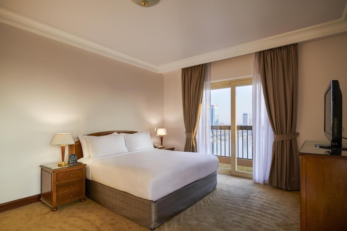 A hotel room with a bed, nightstand, lamp, TV, and a view of the city through the window.