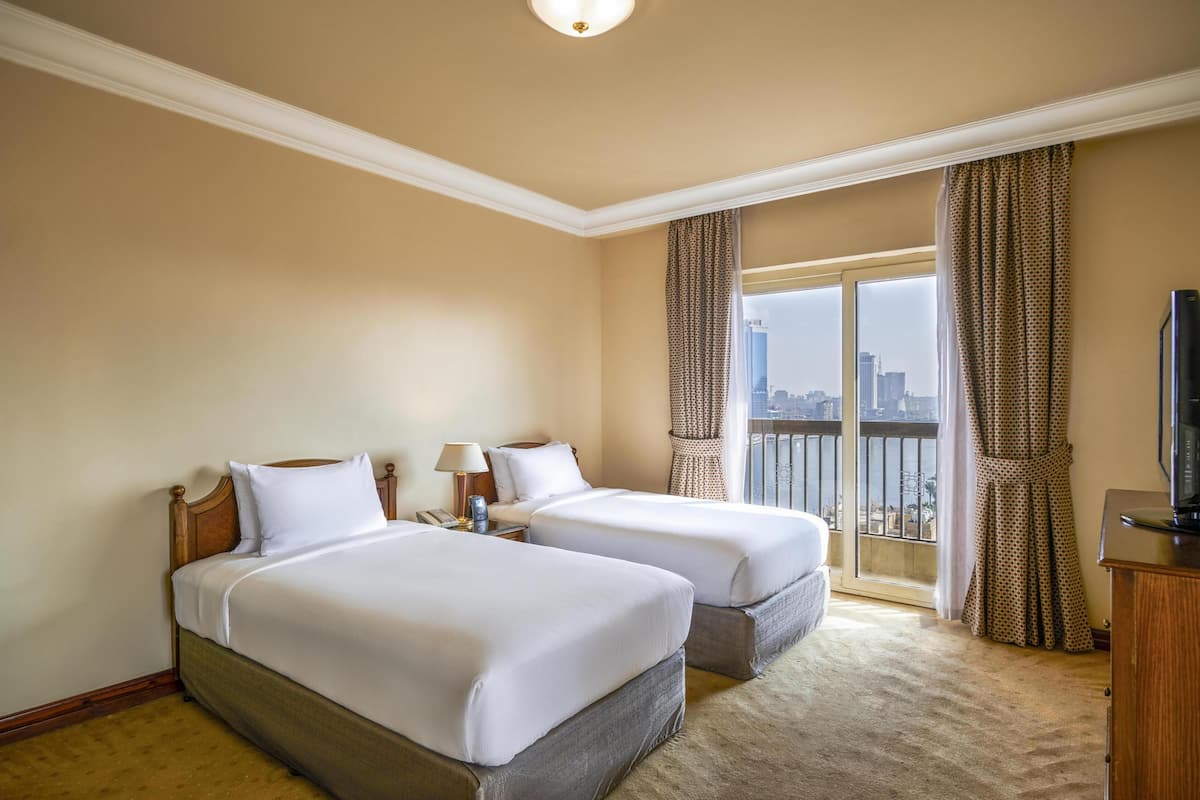 A hotel room with two beds, a television, a desk, and a view of the city.