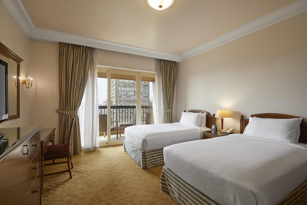 A hotel room with two beds, a desk, a chair, a TV, a lamp, and a view of a building through the window.