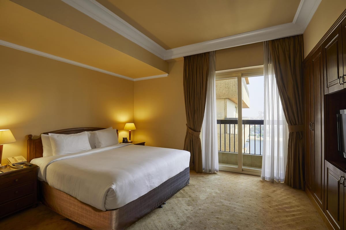 A hotel room with a large bed, a nightstand with a lamp, a TV, and a view of the outside.