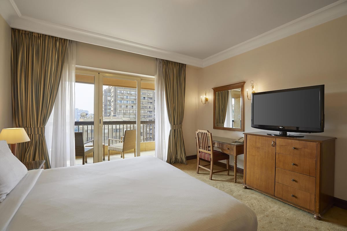 A hotel room with a large bed, a desk with a chair, a television, and a balcony with curtains.