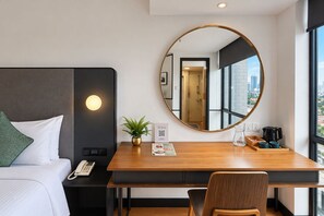 A hotel room with a bed, a desk with a phone, a mirror, a plant, and a window with a city view. - YWCA Fort Canning (Singapore)