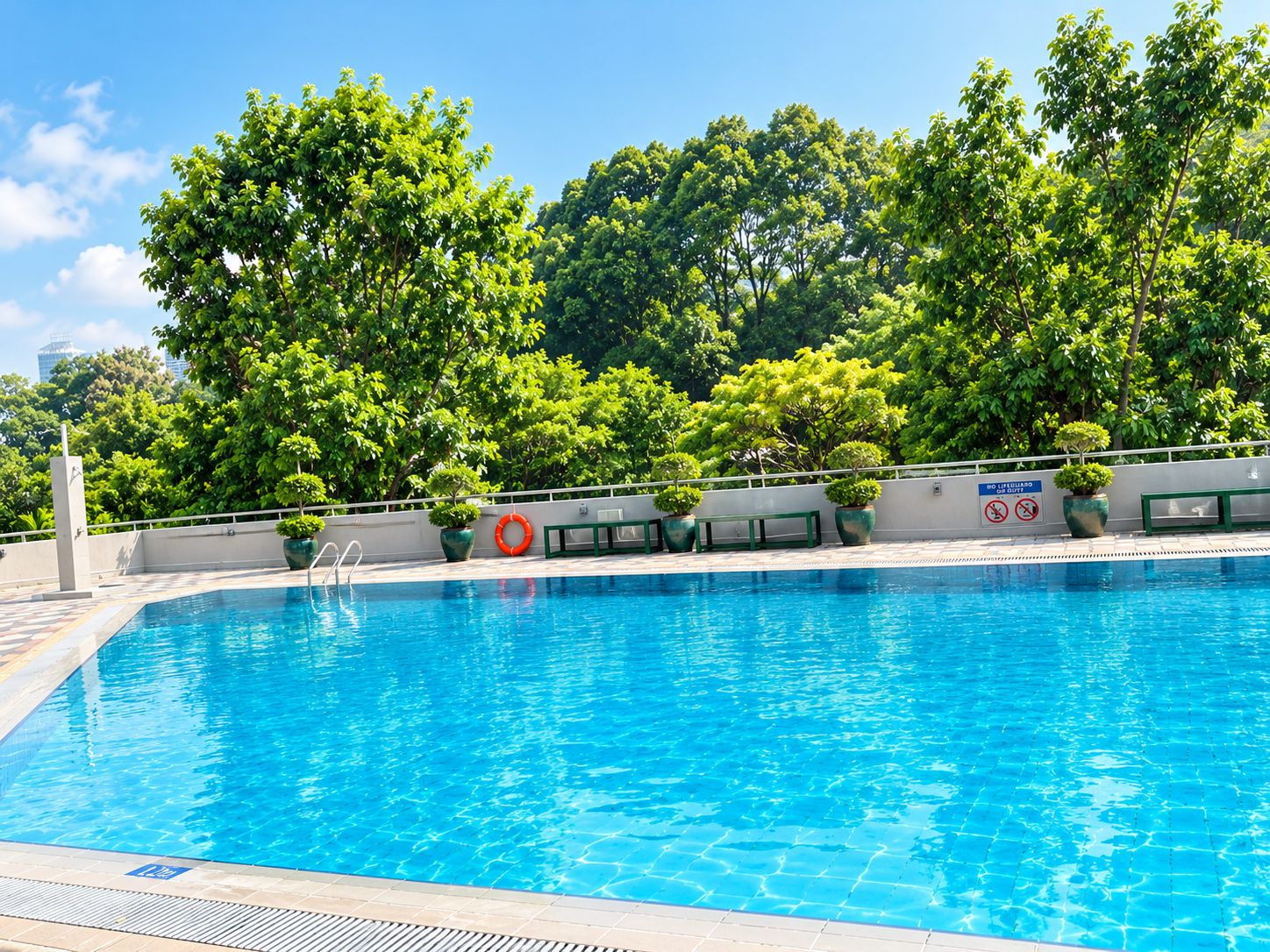 A clear blue swimming pool with a safety sign, surrounded by lush green trees and a white wall with potted plants.