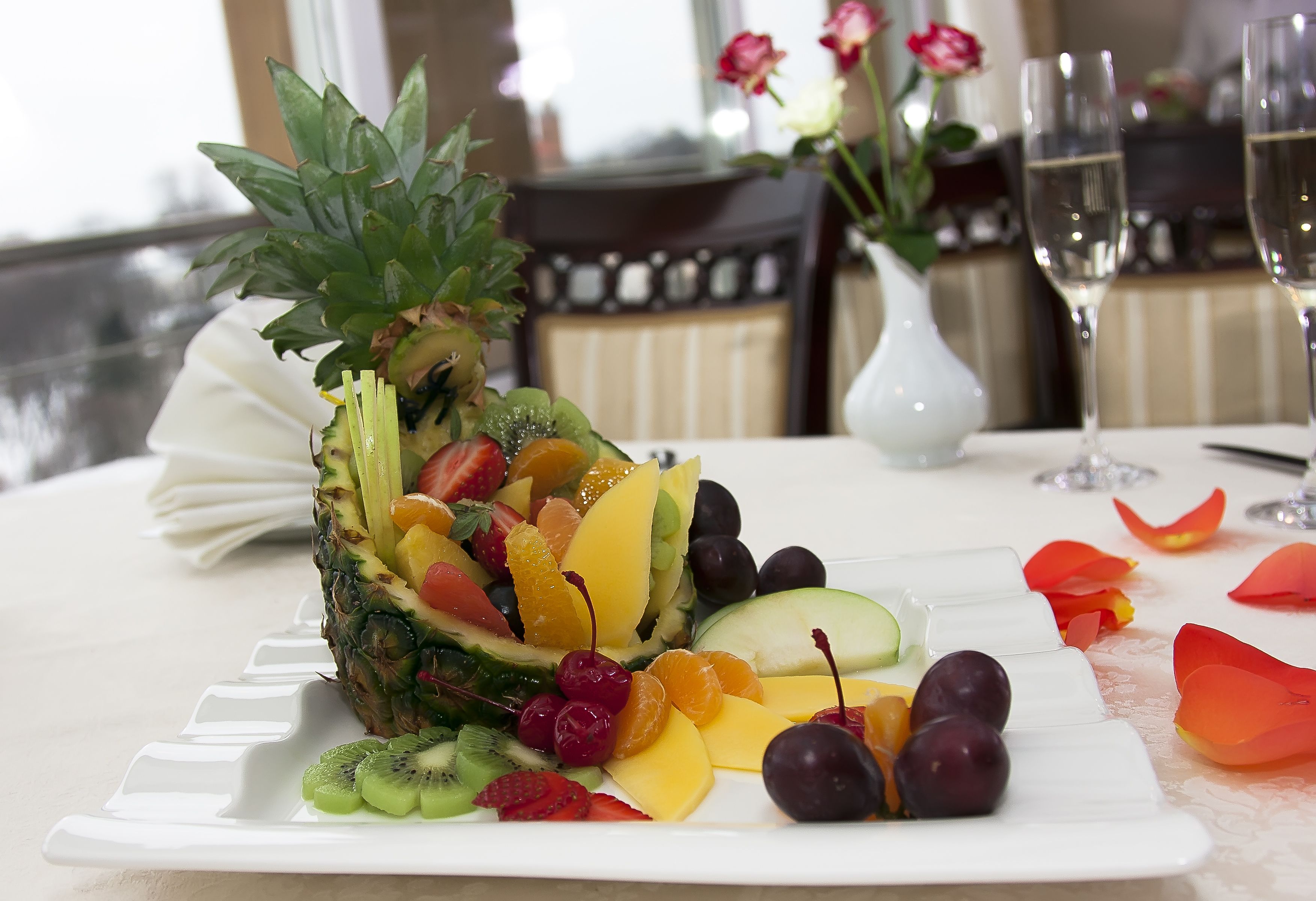 A fruit platter on a white plate with a table setting that includes a vase with flowers and wine glasses.