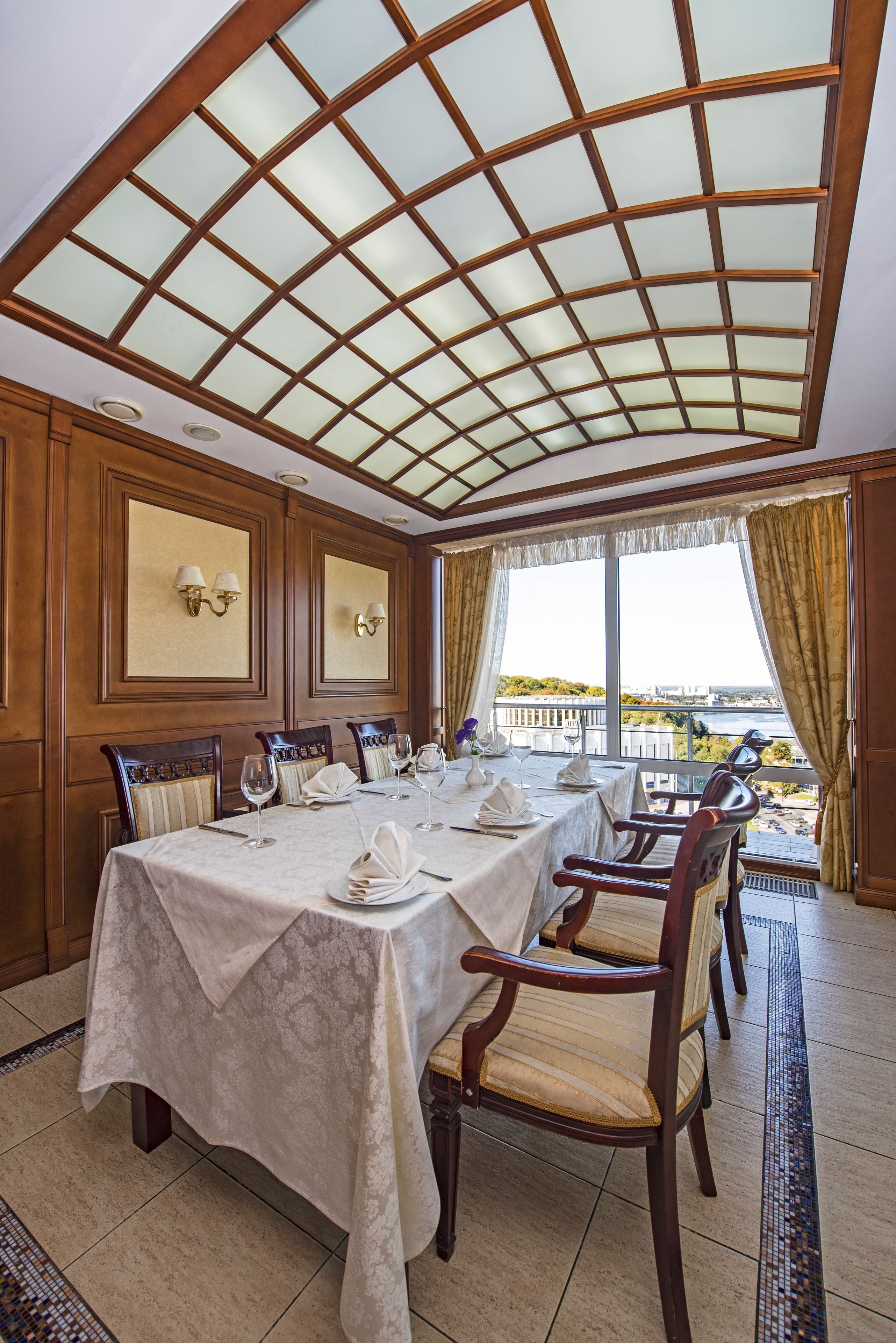 A dining room with a large table set for six, wooden chairs, a glass ceiling, and a view of a body of water.