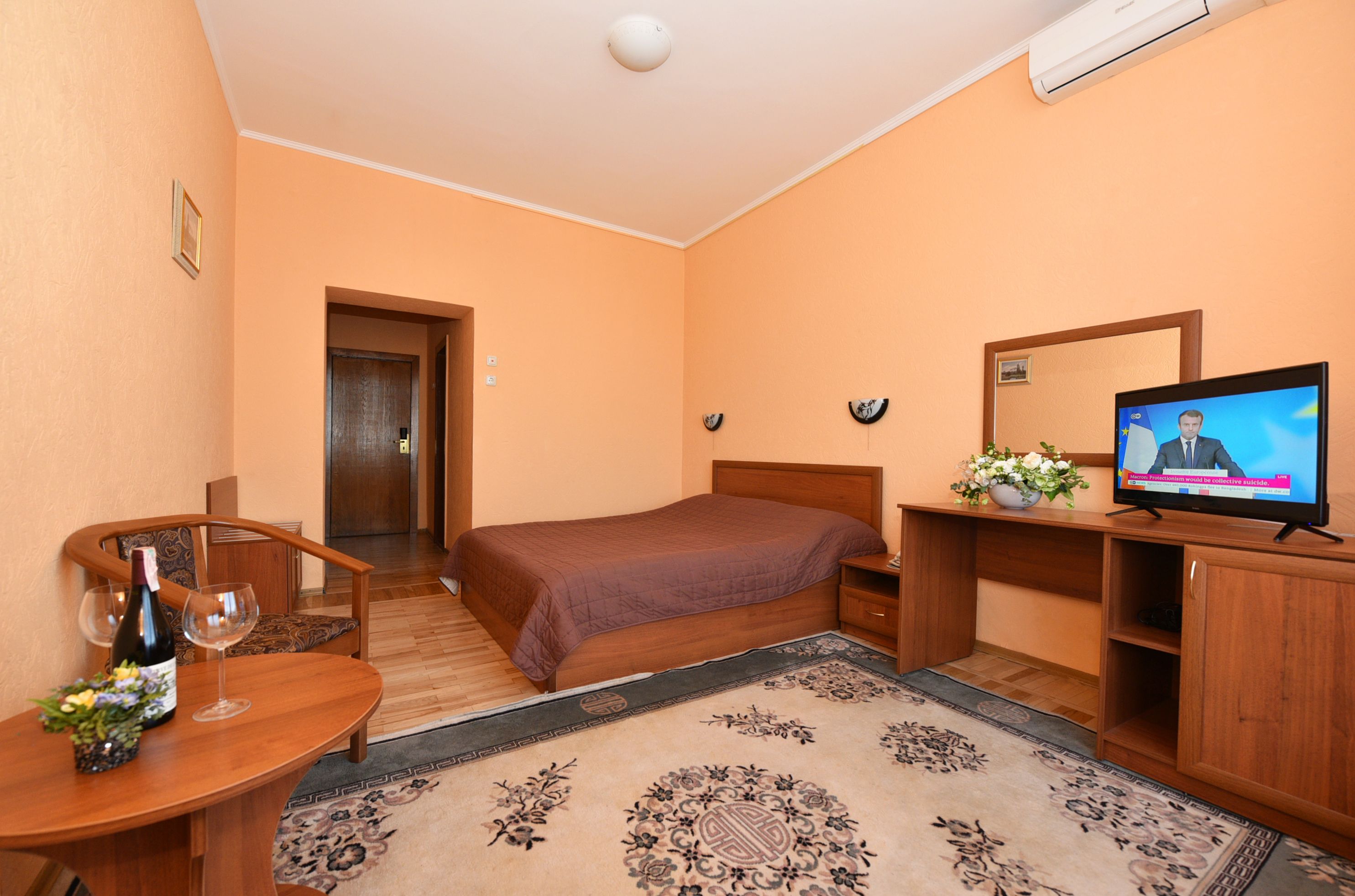 Een hotelkamer met een bed, een bureau met een televisie, een houten stoel, een klein tafeltje met een fles en glazen, en een decoratief vloerkleed.