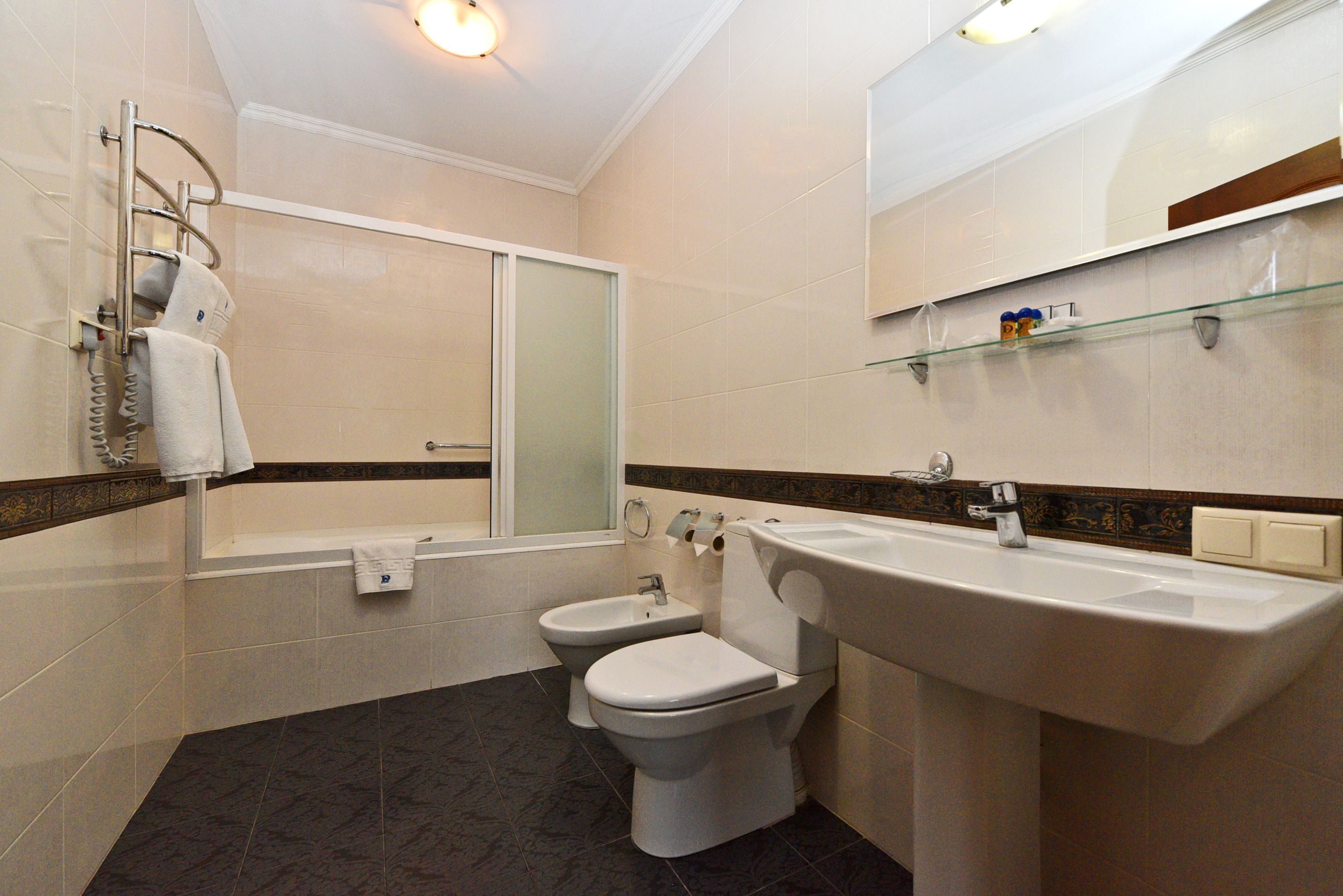 Een moderne badkamer met een badkuip, twee toiletten, een grote wastafel en een glazen plank.