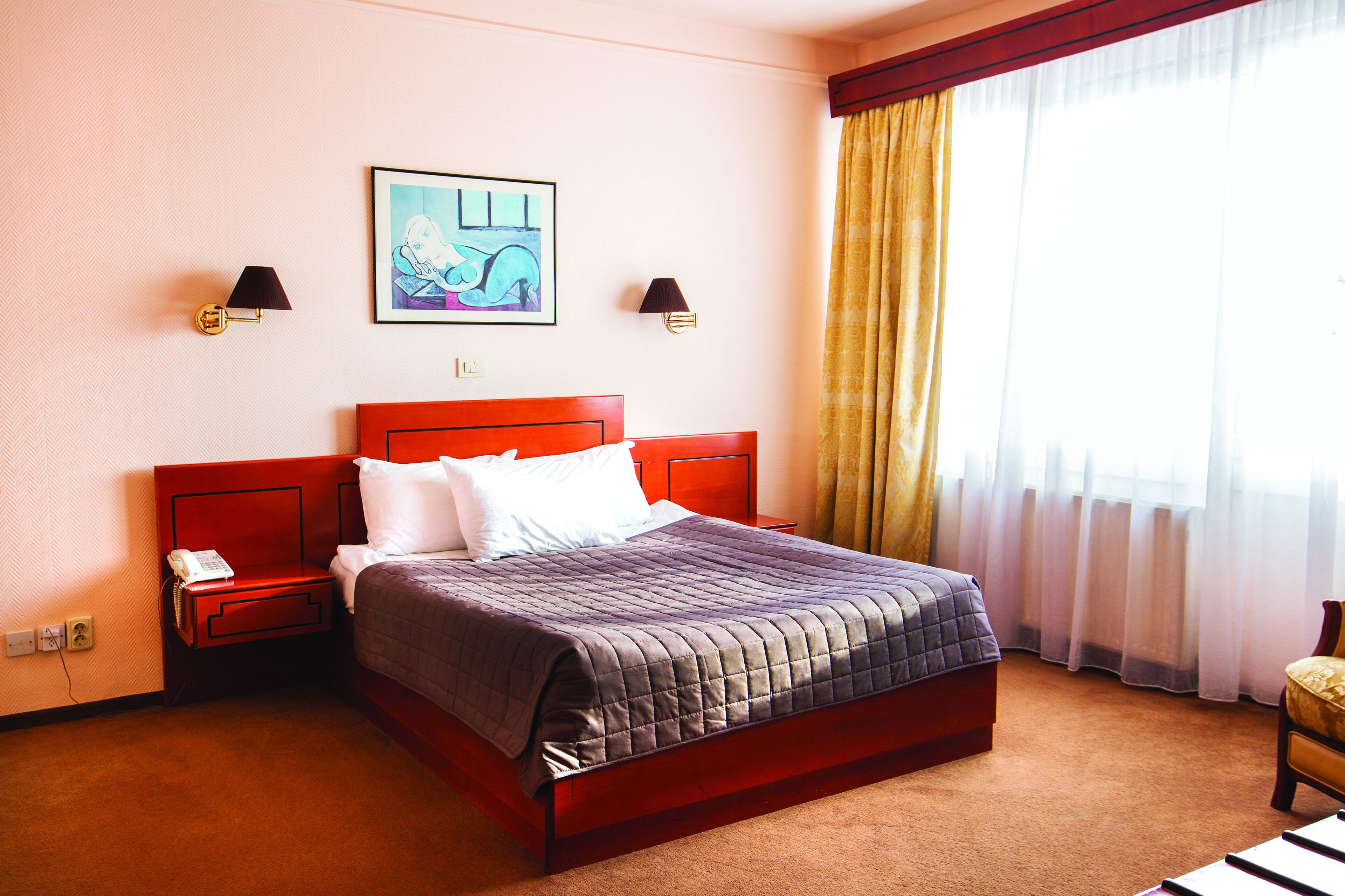 Una habitación de hotel con una cama grande, dos mesitas de noche, una lámpara de noche, un cuadro en la pared y una ventana con cortinas.
