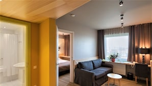 A hotel room with a wooden ceiling, a grey sofa, a small round table, a desk with a lamp, a window with curtains, and a view of the city. - Ibis Styles Riga (Riga)