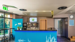 A reception area with a blue counter, a digital screen, and a bar area with shelves of bottles and glasses. - Ibis Styles Riga (Riga)