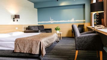 A hotel room with a bed, a desk with a lamp, a chair, a small table with a plant, and a cityscape mural on the wall.
