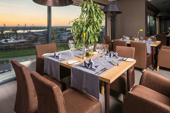 A dining area with tables set for six, large windows offering a view of the cityscape, and a potted plant in the center. - Ibis Styles Riga (Riga)