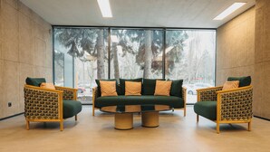 A modern lounge area with a sofa and two wicker chairs, a round coffee table, and large windows offering a view of trees and a parking lot. - Mimi Hotel Chisinau (Chisinau)