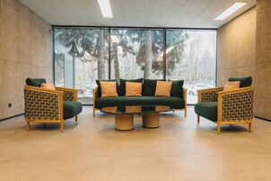Un área de descanso moderna con un sofá y dos sillones de mimbre, una mesa redonda y ventanas amplias que ofrecen vistas a árboles y un estacionamiento.