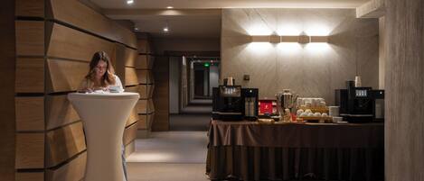 A modern hotel corridor with a reception desk, a buffet area with coffee machines, and a wooden wall design.