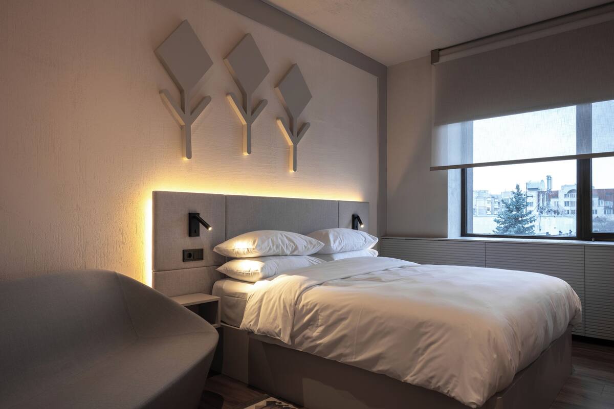 A modern bedroom with a bed, bedside tables, and a window with a city view.