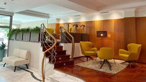 Een moderne lobby met een houten receptiebalie, een trap met glazen balustrade en gele stoelen.