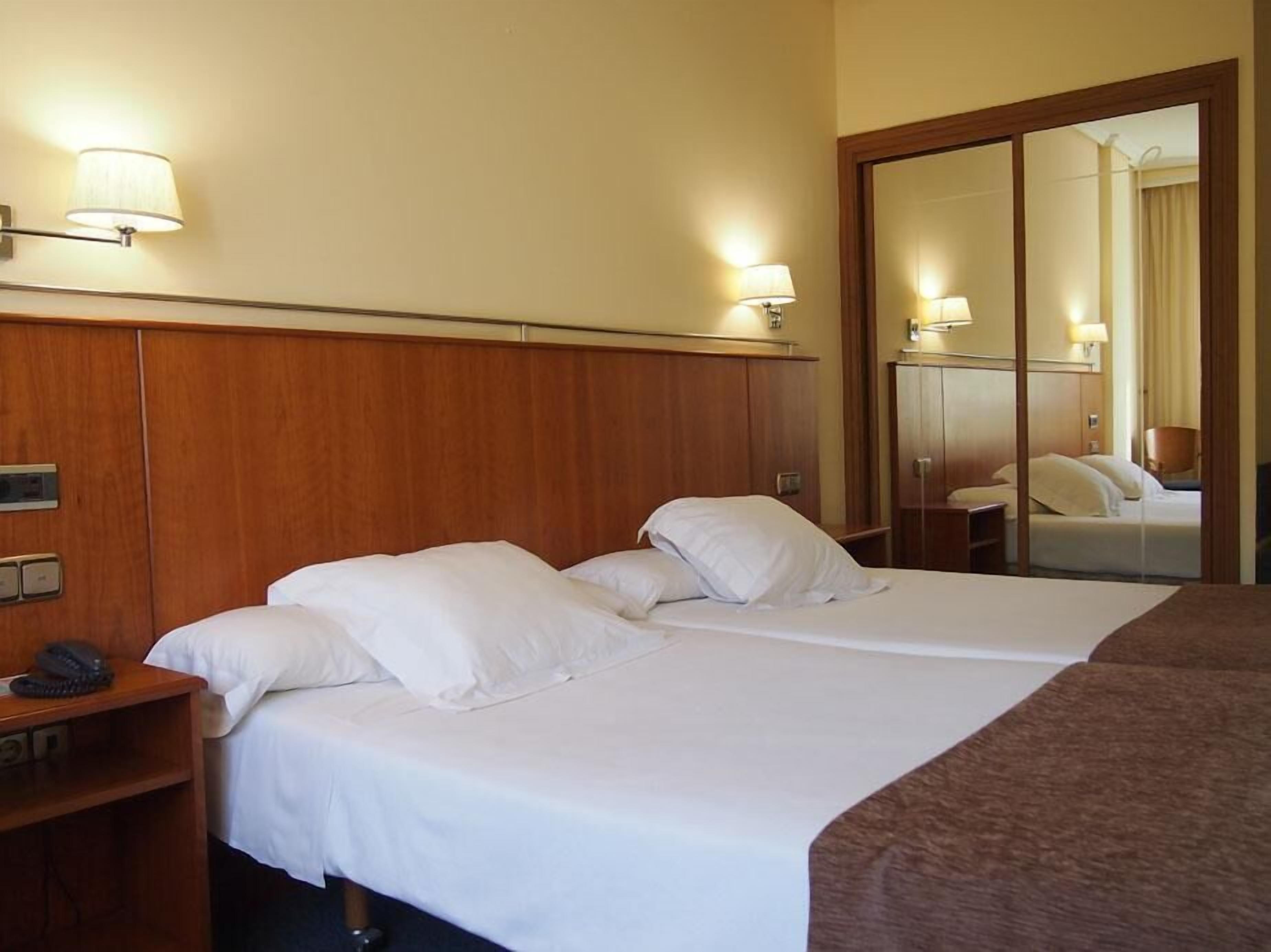 Una habitación de hotel con dos camas, cabecero de madera, lámparas de noche, un teléfono y un espejo.