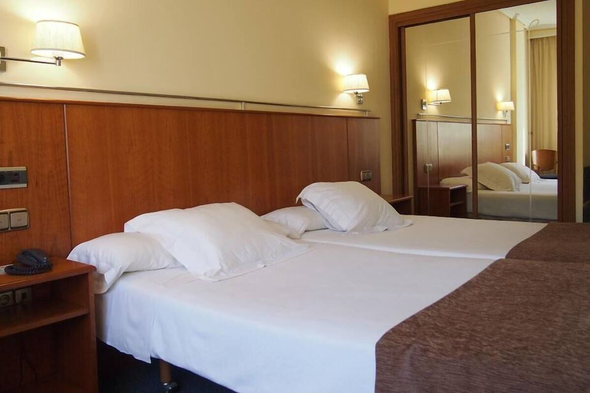 Una habitación de hotel con dos camas, cabecero de madera, lámparas de noche, un teléfono y un espejo.