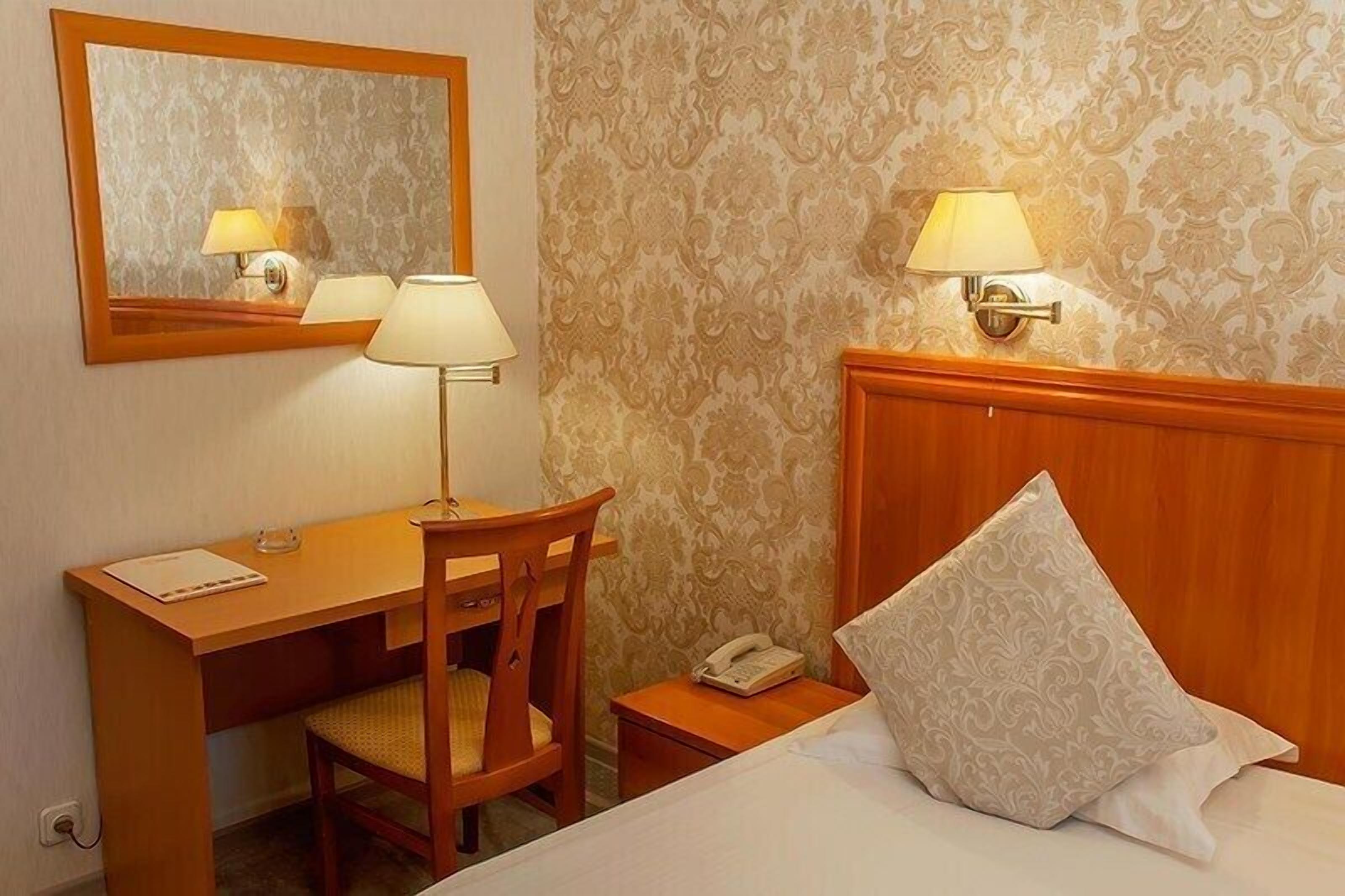 Et hotelværelse med skrivebord i træ, to lamper, en telefon og en seng med et mønstret pudebetræk.