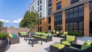 An outdoor seating area with wicker furniture and green cushions, a large planter, and a building with large windows and brick walls. - Hampton Inn & Suites Valley Forge/Oaks (Phoenixville)