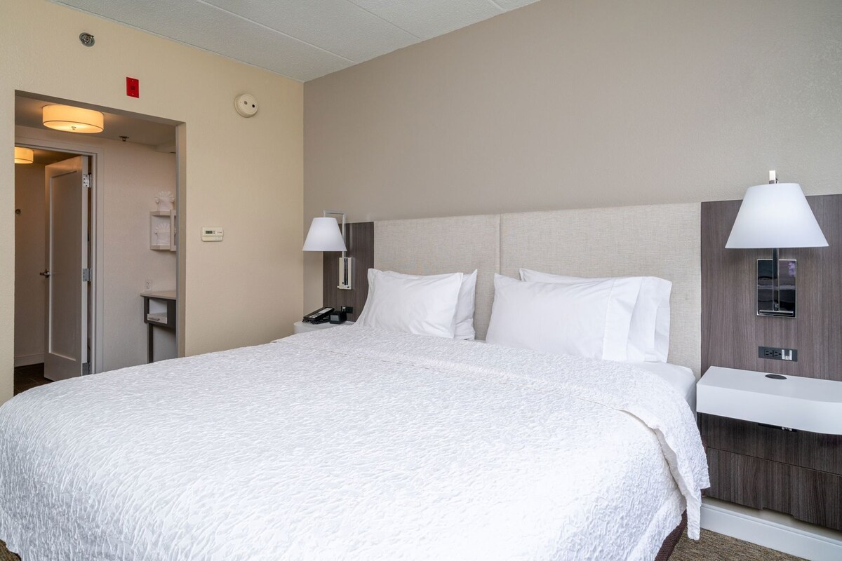 A hotel room with a large bed, bedside lamps, a nightstand, a mirror, and a wall-mounted control panel.