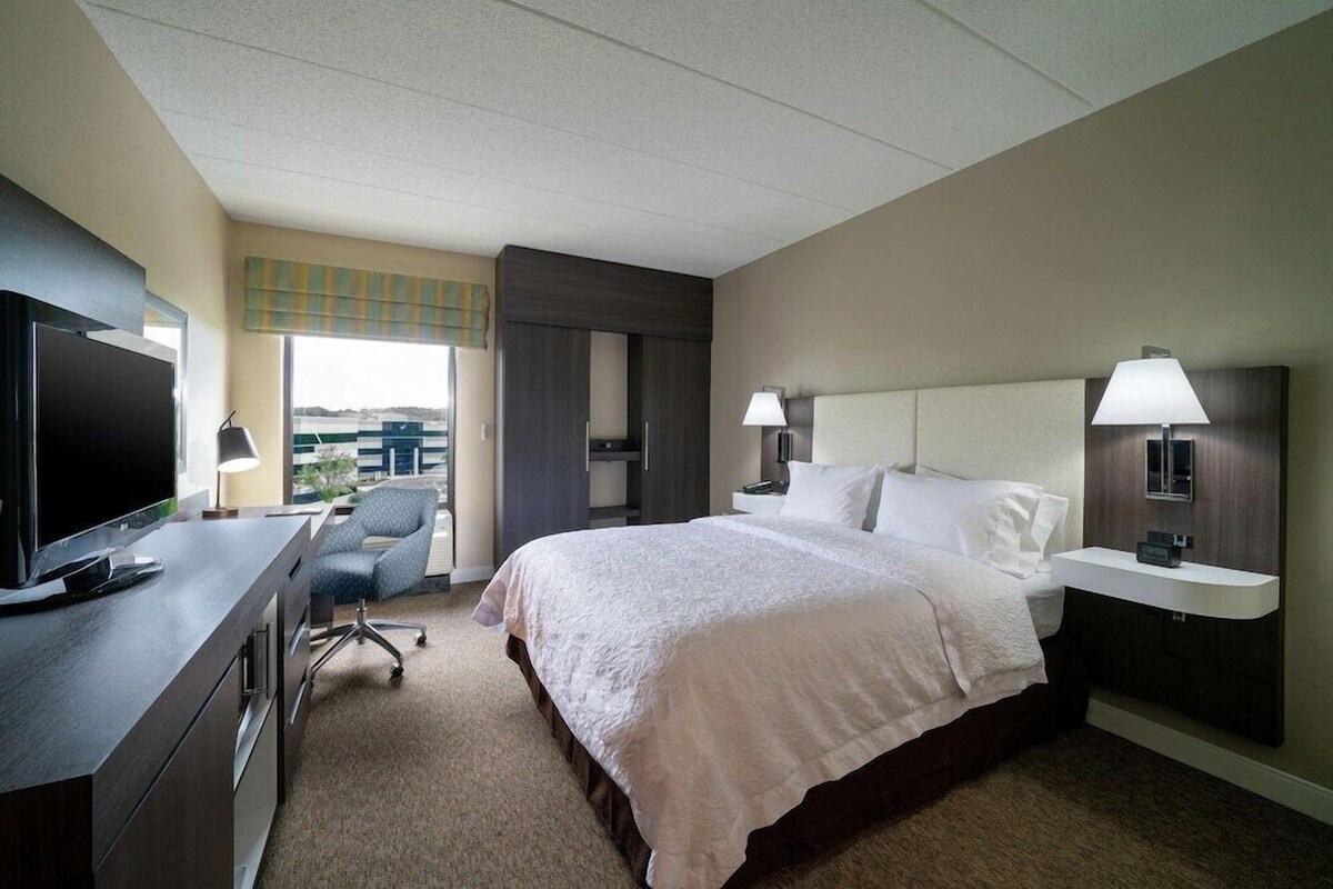 A hotel room with a bed, a desk with a TV, a chair, a bedside table with lamps, and a window with a view.