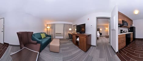 A modern hotel room with a curved layout, a seating area with a sofa and ottoman, a television, a kitchenette, and a separate bathroom area.