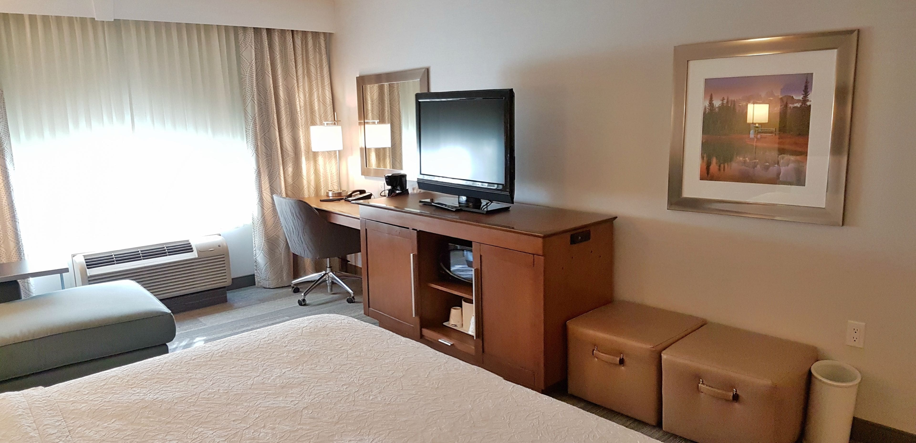Una camera d'albergo con un letto, una scrivania con un computer, una sedia, una televisione, una lampada, un quadro appeso al muro e una finestra con tende.