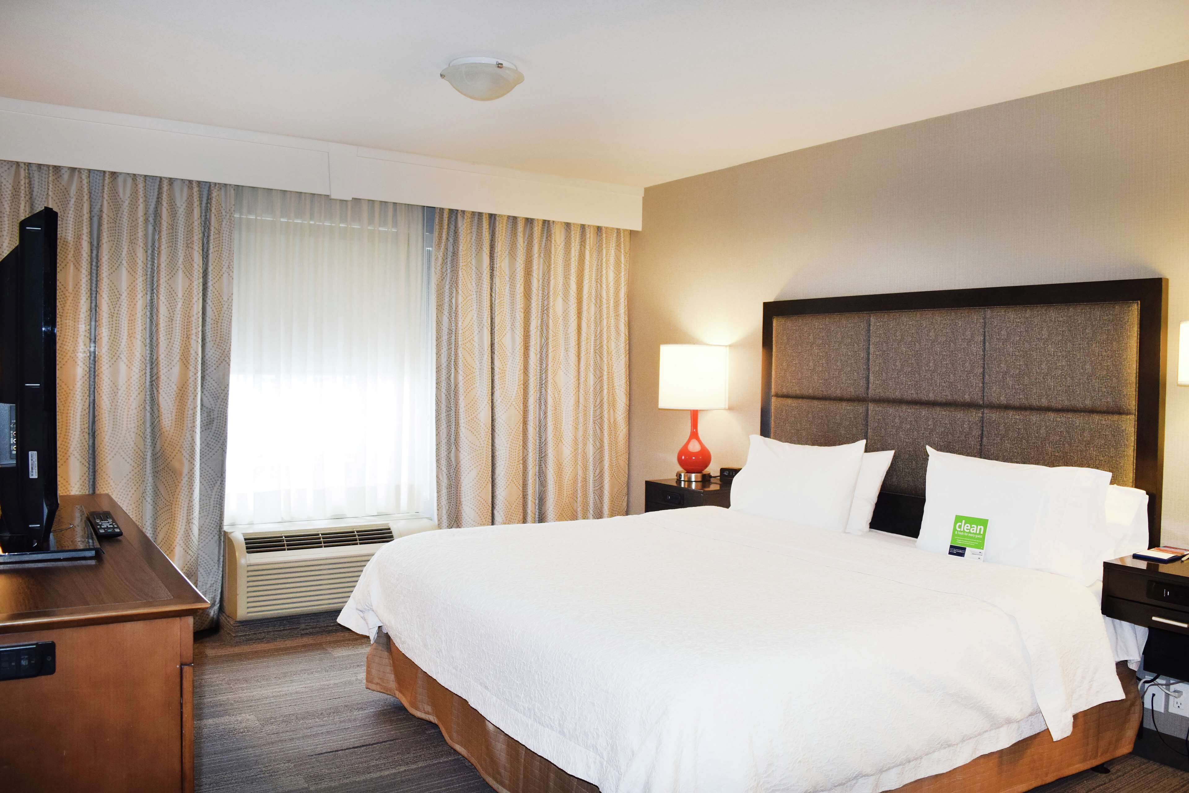 Ein Hotelzimmer mit einem großen Bett, einem Schreibtisch, einer Lampe und Blick ins Freie durch Vorhänge.