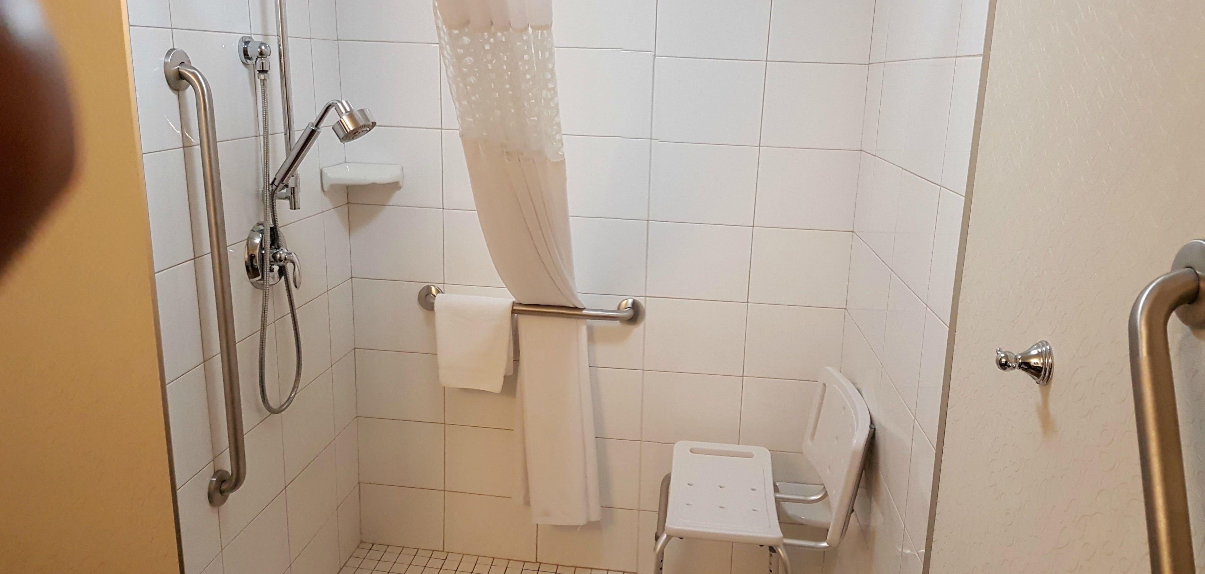 A bathroom with a shower, grab bars, a white toilet, and a small white chair.