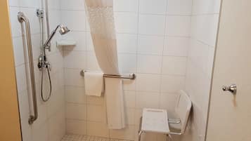 A bathroom with a shower, grab bars, a white toilet, and a small white chair.