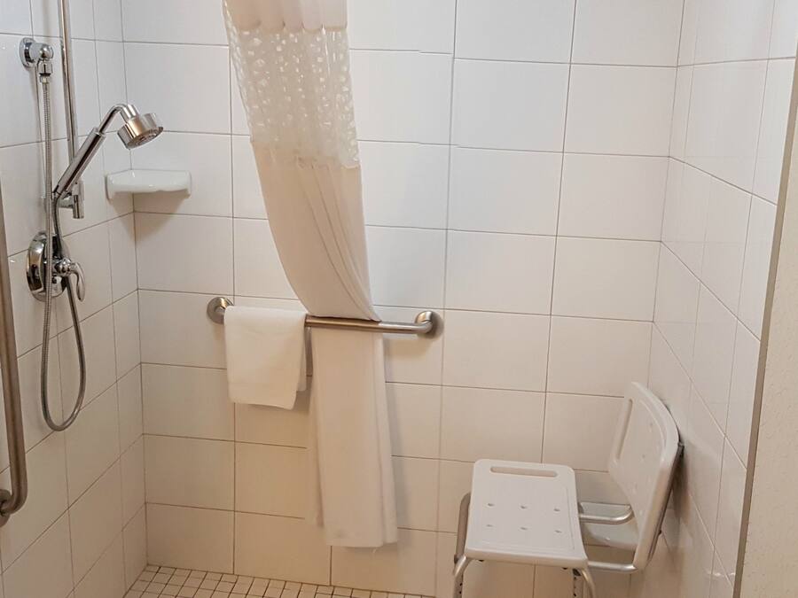 A bathroom with a shower, grab bars, a white toilet, and a small white chair.