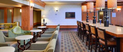 Un vestíbulo de hotel con zona de bar, espacios de asiento y una pantalla de televisión.